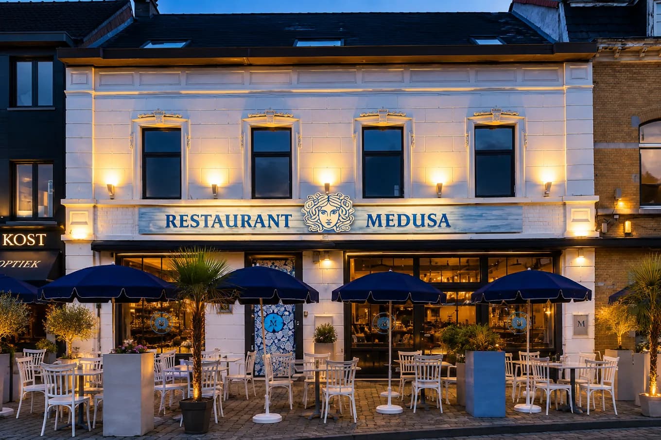 Verlichte witte gevel van Restaurant MEDUSA aan het Heldenplein in Zaventem bij valavond, met terras en blauwe parasols